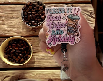 May include: A pink and blue badge holder with a cartoon iced coffee and the text "Fueled by Iced Coffee and Anxiety".