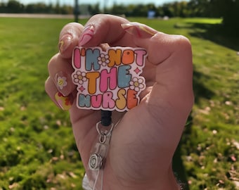 Könnte beinhalten: Ein rosa, blauer, gelber und weißer Badgehalter mit dem Text "I'm not the nurse" in einer Retro-Schriftart. Der Badgehalter ist an einer schwarzen, einziehbaren Badge-Rolle befestigt.