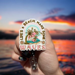 May include: A white, round badge holder with a colourful rainbow and a skeleton surfing on a wave. The badge holder has the text "Life's a Beach, Make Some Waves" in gold lettering.