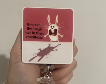 May include: A pink and white badge holder with a cartoon bunny with the text "How can I live laugh love in these conditions".