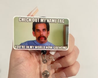 May include: A white plastic badge holder with a printed image of a man with a mustache. The image has the text "CHECK OUT MY NAME TAG" and "YOU'RE IN MY WORLD NOW GRANDMA".