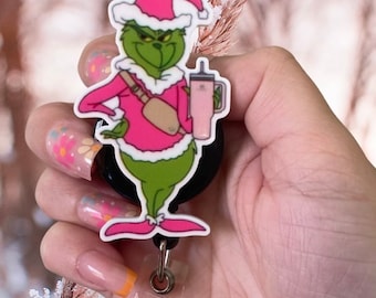 May include: A green Grinch character badge reel wearing a pink Santa hat, sweater, and fanny pack, holding a pink tumbler. The badge reel has a black base and a clear plastic clip.