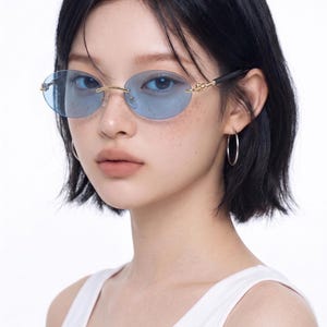 May include: Oval-shaped sunglasses with light blue lenses and gold-colored frames. The sunglasses have a decorative design on the temples and are worn by a person with dark hair and a white tank top.