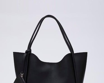 May include: A black tote bag with two shoulder straps, a curved top opening, and a vertical seam down the center. A small, rectangular black pouch is attached to one of the straps by a thin cord. A small rectangular label is on the bottom right.
