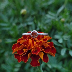 May include: A silver ring with a round orange gemstone set in the centre, resting on a red and orange marigold flower.