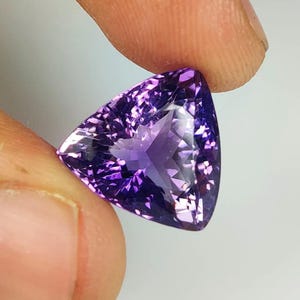 Puede incluir: Una gema triangular facetada en tonos morados. La gema se sostiene entre dos dedos, mostrando su talla y color. Las facetas crean un patrón en forma de estrella en el centro, reflejando la luz. Esta gema es probablemente para la fabricación de joyas.