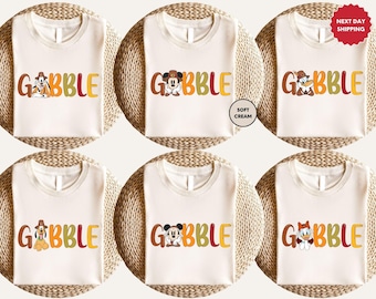 May include: Cream-colored t-shirts with the word "GOBBLE" in brown, orange, green, and yellow. Each shirt features a different cartoon character. The shirts are displayed on woven mats.