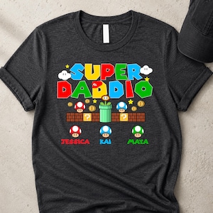 May include: Dark grey t-shirt featuring a colourful "SUPER DADDIO" graphic in a video game style. The design includes mushrooms, coins, and the names Jessica, Kai, and Maya.