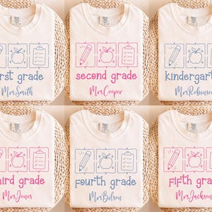 May include: Six off-white t-shirts featuring school-themed graphics including a pencil, apple, and clipboard in squares. Each shirt displays a grade level from kindergarten to fifth grade and a teacher's name in blue or pink script lettering.