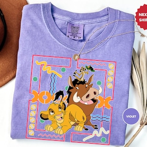 May include: Purple t-shirt featuring a graphic of Simba, Pumbaa, and Timon from The Lion King. The graphic is in a 90s style with a colourful geometric background. The shirt is made of a soft, comfortable material.