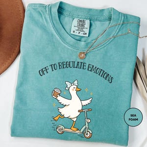 May include: Sea foam green t-shirt with the phrase "OFF TO REGULATE EMOTIONS" and a cartoon duck riding a scooter while holding a coffee cup. The duck has a blue bow and the shirt has a Comfort Colors tag.