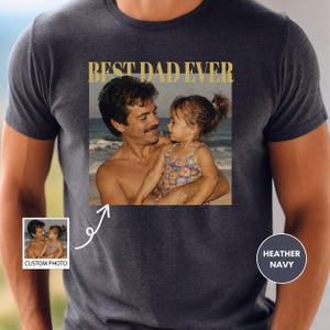 May include: Heather navy t-shirt featuring a photo of a father and child on a beach, with the gold text "BEST DAD EVER". A smaller photo with the text "CUSTOM PHOTO" is also visible.