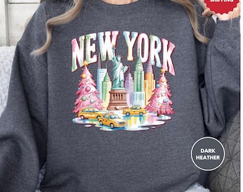 May include: Dark heather gray sweatshirt featuring a colorful illustration of New York City. The design includes the Statue of Liberty, skyscrapers, yellow taxis, and Christmas trees, with the text "NEW YORK" across the top.