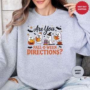May include: A light grey sweatshirt with a Halloween-themed graphic. The design features four cartoon ghosts reading books, pumpkins, bats, and the text "Are You Fall-O-Ween Directions?"