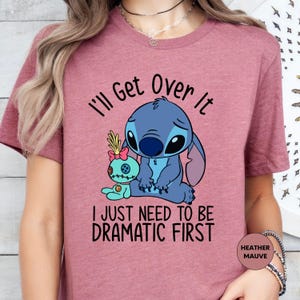 A pink t-shirt with a graphic design featuring a cartoon character and text that reads "I'll Get Over It" and "I JUST NEED TO BE DRAMATIC FIRST".