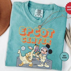 May include: A teal blue t-shirt with a retro-style graphic of Mickey Mouse and a purple alien character. The graphic says "EPCOT CENTER" in orange and yellow letters. The shirt is made of a soft, comfortable fabric and has a distressed look.