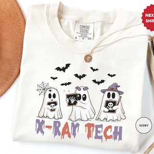 May include: Cream-coloured t-shirt with a Halloween-themed design. Three ghost characters are depicted, each holding an X-ray. The text "X-RAY TECH" is printed below. The design also includes bats and a spiderweb.