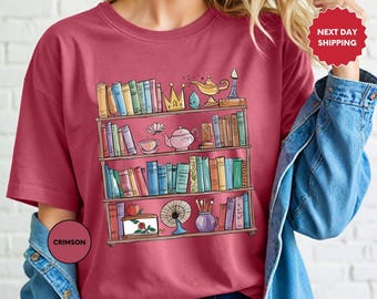 May include: A crimson t-shirt with a graphic of a bookshelf filled with books, a teapot, a crown, a magic lamp, a flower, a cake, and paintbrushes. The graphic is in a cartoon style.