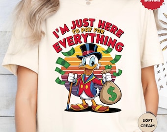May include: Cream-colored t-shirt featuring a cartoon duck in a top hat and suit, holding a money bag and cane. The shirt has the text "I'M JUST HERE TO PAY FOR EVERYTHING" with dollar bills around the duck. The design is set against a sunset.