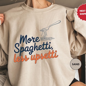 May include: A beige sweatshirt with a graphic of a fork with spaghetti on it. The text on the sweatshirt says "More Spaghetti, Less Upsetti".