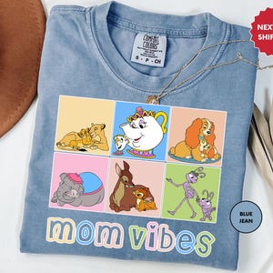 May include: A light blue t-shirt featuring a graphic of Disney characters and the text "mom vibes" in colourful letters. The characters include Belle, Lady, Simba, Dumbo, and Flik.