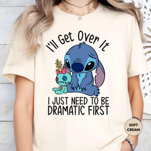 A white t-shirt with a graphic design featuring a blue cartoon character and text that reads "I'll Get Over It" and "I just need to be dramatic first".