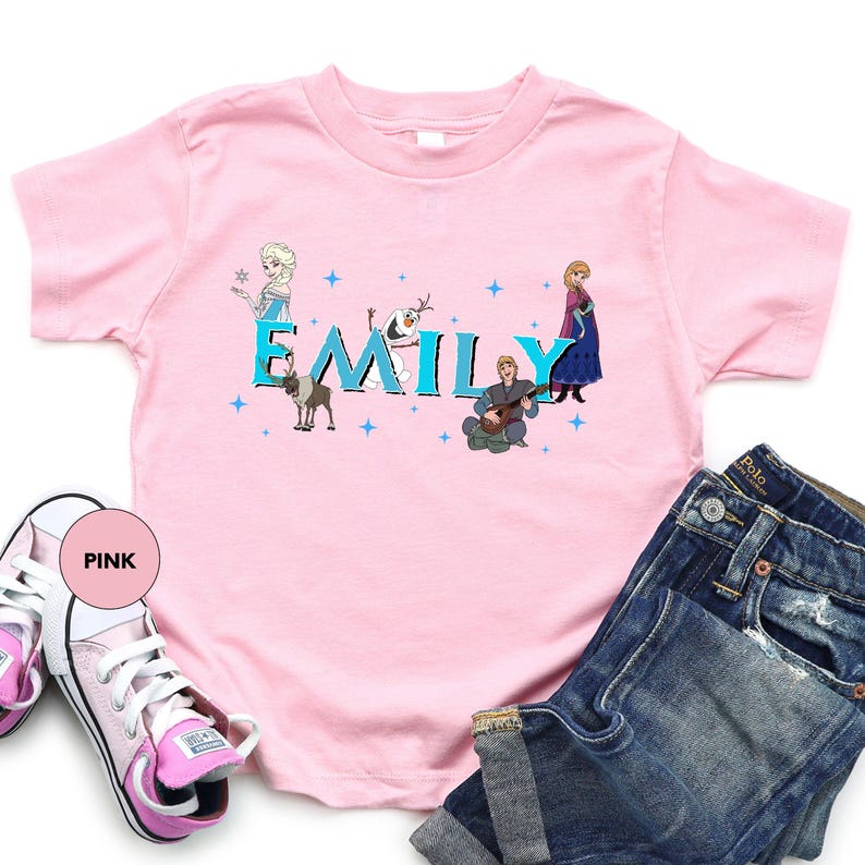 A pink t-shirt with the name "EMILY" printed on it, along with illustrations of characters from the Disney movie Frozen. The t-shirt is placed on a white background, and there are also a pair of jeans and pink sneakers visible in the image.