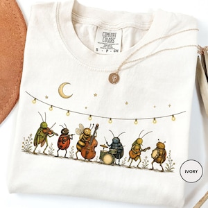 May include: An ivory t-shirt featuring a graphic of six insects playing musical instruments under a string of lights and a crescent moon. The insects include a grasshopper with a violin, a ladybug with a flute, a bee with a bass, a beetle on drums, a grasshopper with a guitar, and a beetle with a tambourine.