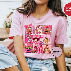 May include: A light pink t-shirt featuring a grid of cartoon character illustrations. The design includes characters like Mr. Potato Head, Buzz Lightyear, and others, with a Valentine's Day theme. The shirt has short sleeves.