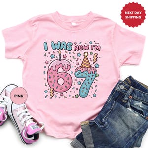 May include: Pink toddler t-shirt with the text "I WAS NOW I'M 67" in colorful letters and numbers. The number 6 is a pink donut with sprinkles and a candle. The number 7 is blue with a party hat.