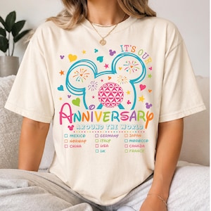May include: A cream-colored t-shirt featuring a colorful graphic of a mouse-ear silhouette containing fireworks and a geodesic sphere. The text reads IT'S OUR ANNIVERSARY AROUND THE WORLD, followed by a checklist of countries including Mexico, Norway, China, Germany, Italy, USA, UK, Japan, Morocco, Canada, and France.