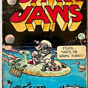 May include: A vintage paperback book cover titled STAR JAWS by Will Eisner. The illustration features a robot in a flying saucer in space, with a speech bubble saying STUPID... THAT'S THE WRONG PLANET! A sign in the foreground reads YOU ARE ENTERING EARTH.