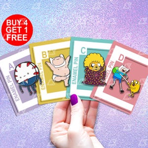 May include: Four enamel pins featuring characters from the animated television series Adventure Time. The pins are labeled A, B, C, and D. The characters are Finn the Human, Jake the Dog, Marceline the Vampire Queen, and Ice King. The pins are on a pink, blue, and purple background.
