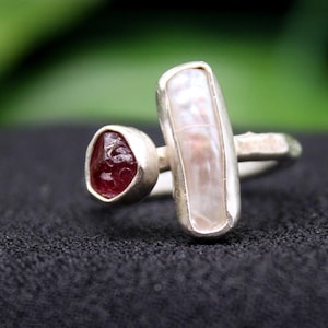 May include: A silver ring featuring two gemstones. One is a small, triangular, deep red stone, and the other is a larger, rectangular, off-white stone. The ring is set against a dark background.