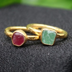 Pode incluir: Duas alianças de ouro com pedras preciosas quadradas. A aliança à esquerda tem uma pedra vermelha, e a aliança à direita tem uma pedra verde.