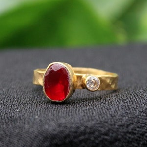 May include: A gold ring with a red oval gemstone and a smaller white gemstone. The ring is on a black fabric background.