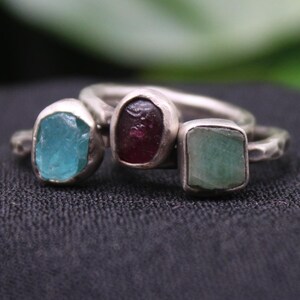 May include: Three silver rings with different colored gemstones. The ring on the left has a light blue gemstone, the middle ring has a dark red gemstone, and the ring on the right has a light green gemstone.