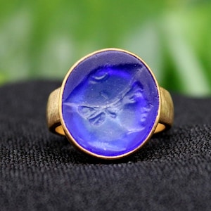 May include: A gold ring with a blue glass cameo featuring a carved design. The cameo is round and set in a gold band.