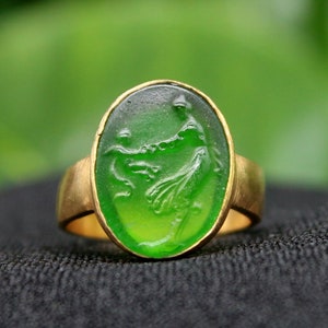Può includere: Un anello d'oro con una gemma verde ovale. La gemma presenta un disegno scolpito di una figura. L'anello è posto su una superficie nera con uno sfondo verde sfocato.