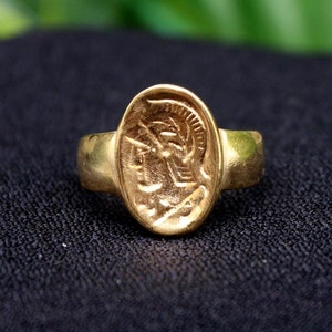 Pode incluir: Um anel de ouro com um design oval que apresenta o perfil de uma pessoa usando um capacete. O anel est&aacute; sobre uma superf&iacute;cie preta.