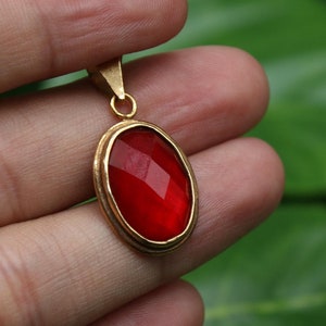 May include: A gold pendant with a red oval-shaped gemstone. The pendant is held in a hand against a green leafy background.