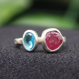 May include: A silver ring featuring two gemstones: an oval, light blue stone and a rounded, deep red stone. The ring is set against a black textured surface with a blurred green background.