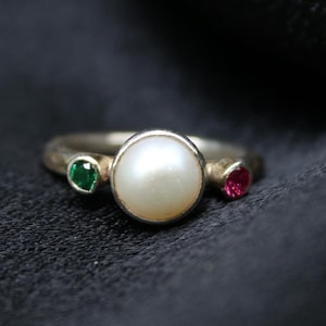 Op de afbeelding: Een zilveren ring met een witte parel, een groene edelsteen en een rode edelsteen.