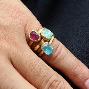 May include: Three gold rings with colorful gemstones. The top ring has a pink gemstone, the middle ring has a light blue gemstone, and the bottom ring has a darker blue gemstone.
