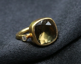 May include: A gold ring with a large, square-cut, olive-green gemstone as the primary focus. A smaller, clear gemstone is set on the band. The ring is displayed on a dark, textured surface.