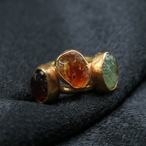 May include: A gold ring with three different colored gemstones. The ring has a dark red, an orange, and a green gemstone.