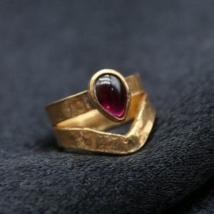 Può includere: Un anello d'oro con una pietra preziosa di colore rosso scuro a forma di lacrima incastonata al centro. L'anello è composto da due bande, una delle quali è una banda semplice e l'altra è una banda a forma di V.