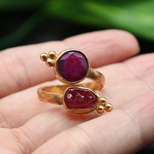 Könnte beinhalten: Ein goldener Ring mit zwei roten Edelsteinen. Der Ring ist so konzipiert, dass er sich um den Finger wickelt.