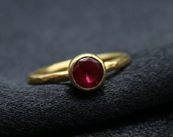 May include: A gold ring with a red gemstone set in the center. The ring is on a black fabric background.