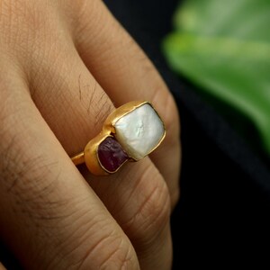 May include: A gold ring featuring a white pearl and two red gemstones. The ring is worn on a finger.